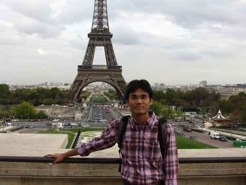 A man in a plaid shirt and jeans leans on a stone balustrade, the Eiffel Tower rising behind him under an overcast sky.