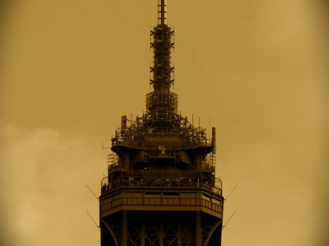 The top platform and antenna of the Eiffel Tower in a sepia-toned, vignetted close-up against a hazy sky.