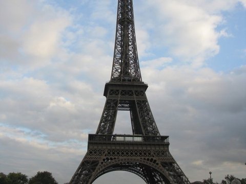 The Eiffel Tower towering over the Pont d'Iéna, a long glass-roofed river boat passing on the Seine beneath scattered cloud.