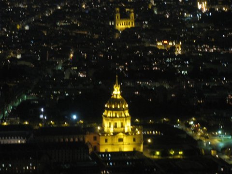 The floodlit golden dome of Les Invalides glowing amid a dark sea of Paris city lights, seen from high above.