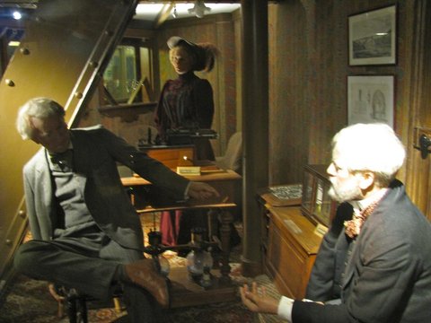 Two wax figures in nineteenth-century suits converse in a small wood-panelled office, a third standing by a desk at the back.