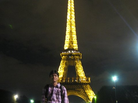 A man in a plaid shirt stands on a dark pavement with the golden-lit Eiffel Tower rising behind him under a night sky.