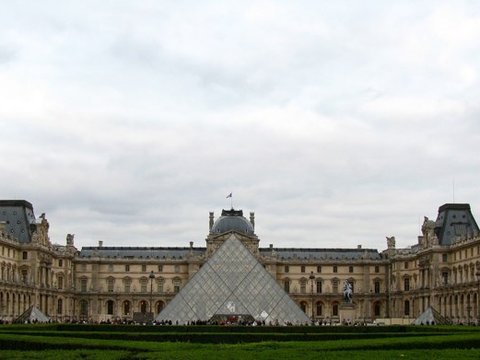 Wide view of the Louvre's long stone wings and slate domes framing the glass pyramid under an overcast sky, visitors tiny at its base.