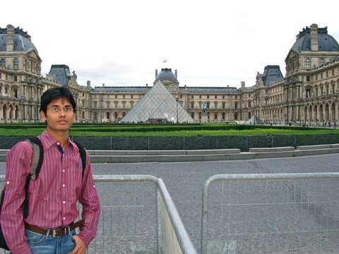 A man in a striped shirt with a backpack stands at a metal barrier, the Louvre's glass pyramid and palace wings behind him.