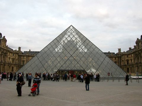The Louvre's glass pyramid fills the frame beneath a grey sky, a crowd of visitors gathered at its base and a pushchair in the foreground.