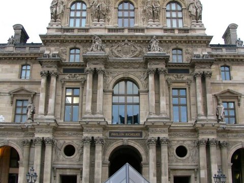 Sculpted stone facade of the Pavillon Richelieu rising above a small glass pyramid, visitors blurred in motion at its foot.
