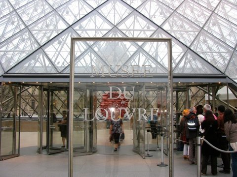 Revolving doors beneath a steel frame lettered Musee du Louvre, the glass pyramid's lattice overhead and visitors queuing at the right.