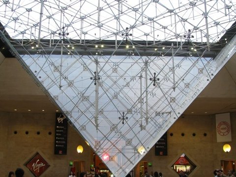 The Louvre's inverted glass pyramid hangs point-down above a small stone pyramid in a bright underground hall, shoppers passing beneath.