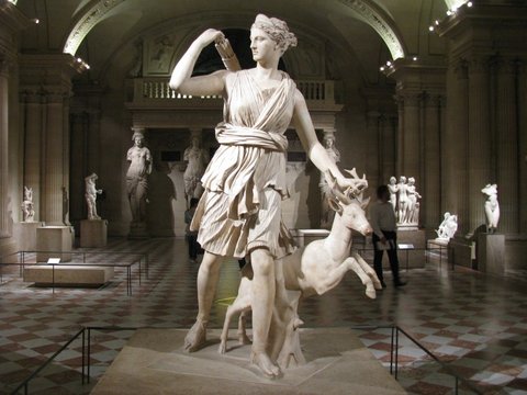 Marble statue of a striding goddess reaching for an arrow with a leaping stag at her side, in a lamplit sculpture gallery.
