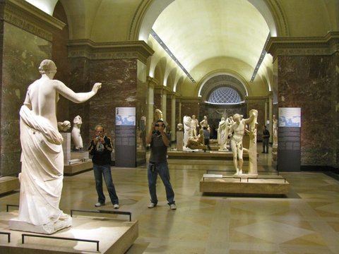 A vaulted gallery of white marble statues on stone plinths, two visitors raising cameras between the red marble pilasters.