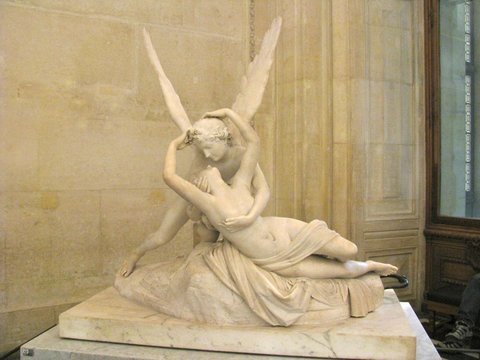 Marble sculpture of a winged figure cradling a reclining woman, carved drapery spilling over the plinth against a pale stone wall.