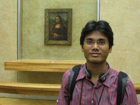 A man in a striped shirt with an audio guide stands close to the camera, the Mona Lisa hanging on the wall behind him.