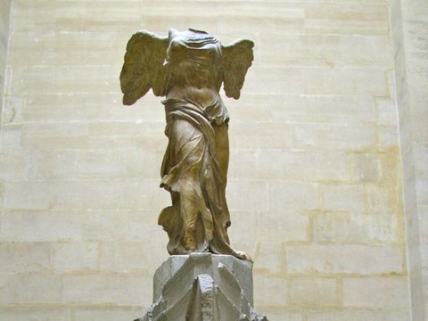 A headless winged marble figure in wind-swept drapery stands on a stone ship's prow against pale limestone walls.