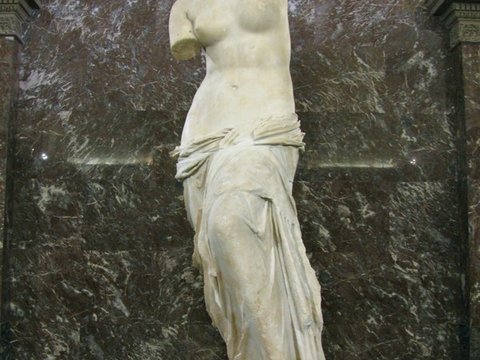 An armless white marble statue of a woman, drapery gathered at her hips, before a dark red marble niche.