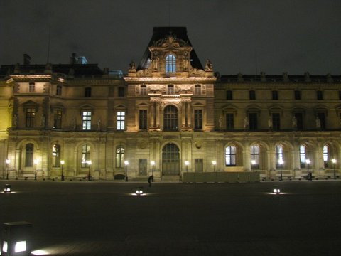 A floodlit stone palace wing with a tall pavilion roof and rows of lit windows above a dark, almost deserted cobbled square.