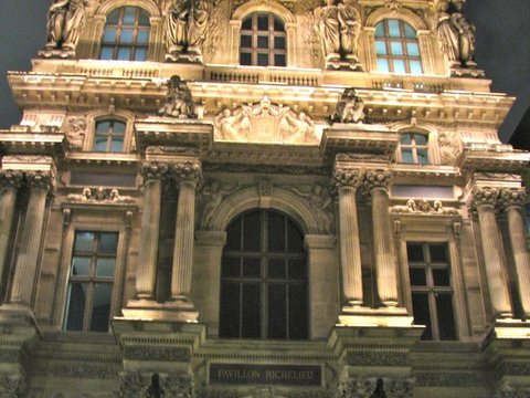 Looking up at a floodlit ornate pavilion with sculpted figures and columns, the tip of a glass pyramid at its foot.