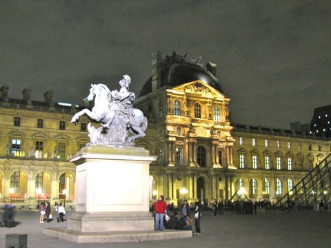 A white stone horse and rider rear on a tall plinth before the floodlit palace facade, visitors gathered below at night.