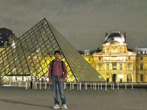 A man in a maroon shirt and jeans stands before the glass pyramid, the floodlit palace glowing behind under a dark sky.