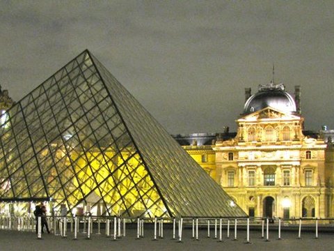 The glass pyramid glows against a dark sky, the floodlit palace wings and domed pavilion stretching behind it.