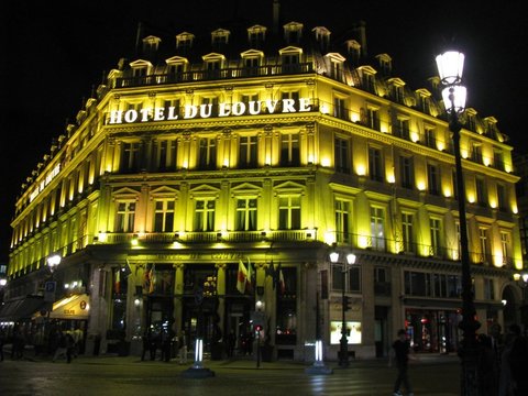 A grand corner building lit golden at night, its name in bright letters above rows of balconied windows and a street lamp.