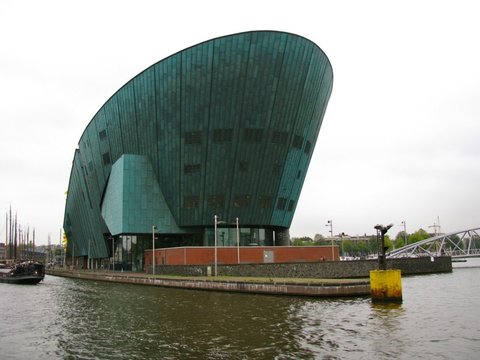 A huge green-copper building shaped like a ship's hull rises from the water beside a brick quay and a yellow mooring post.