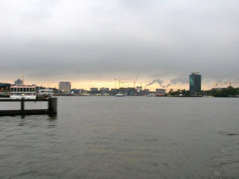 A low city skyline with cranes and a lone tower beneath heavy grey cloud, a pale band of light along the horizon over rippled water.
