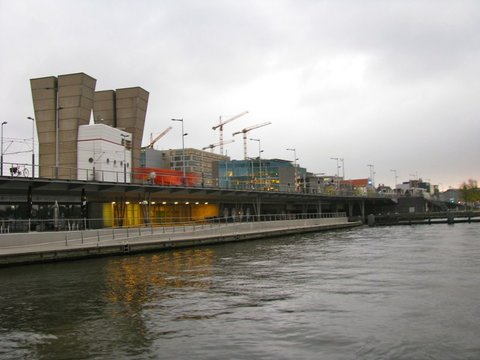 Concrete towers and tall construction cranes rise behind a long glass-fronted quay building on grey water under an overcast sky.