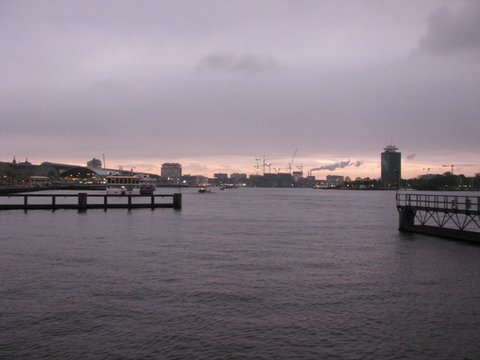 Grey water and wooden jetties lead toward a distant skyline of cranes and a tower, a faint glow of sunset under low cloud.