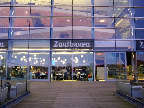 A glass-fronted restaurant glows at dusk, its string of round lamps and seated diners visible behind the Zouthaven sign on a wooden deck.