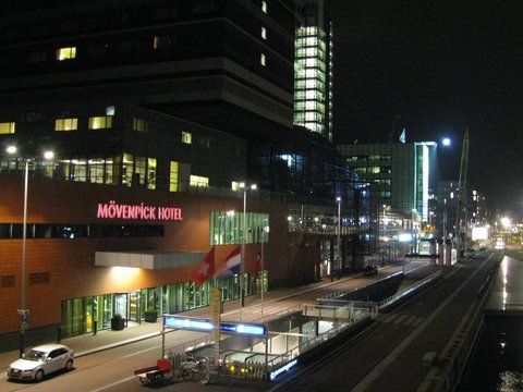A brick hotel with a red neon sign and glowing lobby stands beside a tram stop and a lit glass tower under a black night sky.