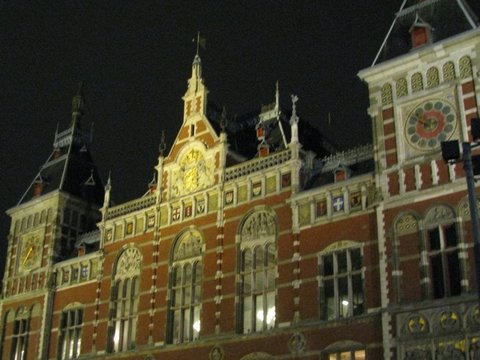 The floodlit red-brick facade of Amsterdam Centraal, with gilded clock, ornamental gables and tall arched windows against a dark sky.