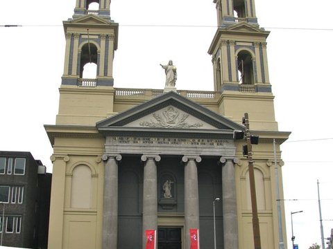 A pale twin-towered church with a columned portico rises over market stalls and parked bicycles beneath a white overcast sky.