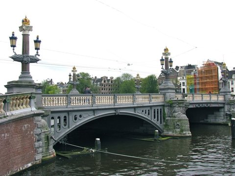 A grey iron bridge with white balustrades and crown-topped lamp posts spans a dark river, canal houses and scaffolding behind it.