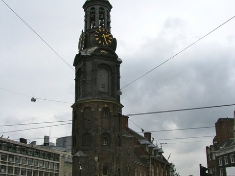 The dark brick Munttoren with its ornate clock and open belfry rises above a busy junction criss-crossed by tram wires under grey cloud.