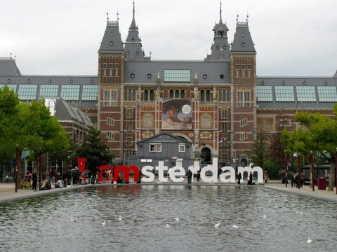 Giant red and white I amsterdam letters stand before the turreted Rijksmuseum, a reflecting pool with gulls in the foreground.