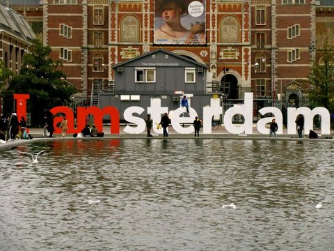 Gulls drift on a rippled pool in front of the I amsterdam letters, a few visitors sitting on them beneath the Rijksmuseum facade.