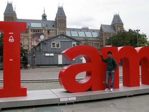 A man in a green hooded top and jeans stands inside the giant red letters of the I amsterdam sign, the Rijksmuseum behind him.