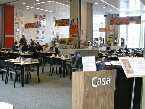 A bright hotel breakfast room with grey carpet, dark chairs at set tables, food posters on the walls and a Casa sign on a wooden stand.