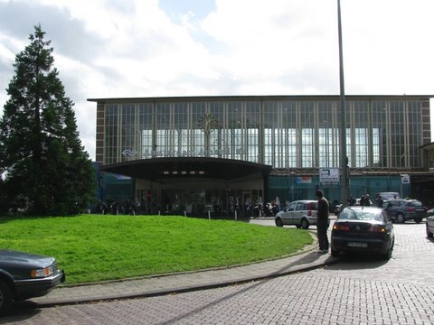 A mid-century station with a tall glass facade and curved canopy faces a green lawn and a tall conifer, cars parked on the cobbled road.