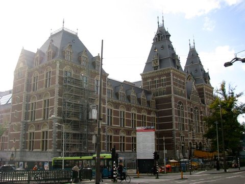 The brick towers and steep slate roofs of the Rijksmuseum, one wing wrapped in scaffolding, above a green bus and cyclists at a junction.