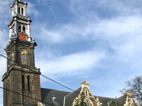 The tall brick tower of the Westerkerk, topped by a blue and gold crown, rises over gabled roofs and street trees beneath a bright, cloud-streaked sky.