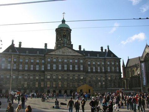 The wide stone front of the Royal Palace with its green-domed cupola, crowds milling across the square below and the Nieuwe Kerk at the right edge.