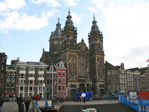 Twin-towered basilica with a central dome and rose window standing among brick houses, a blue van and rows of parked bicycles at its foot.