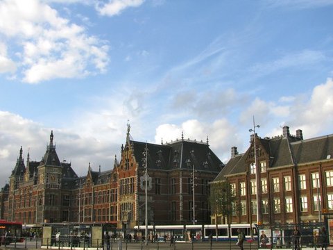 The long red-brick facade of Amsterdam Centraal with its turrets under a bright blue sky scattered with white clouds, trams and buses passing in front.