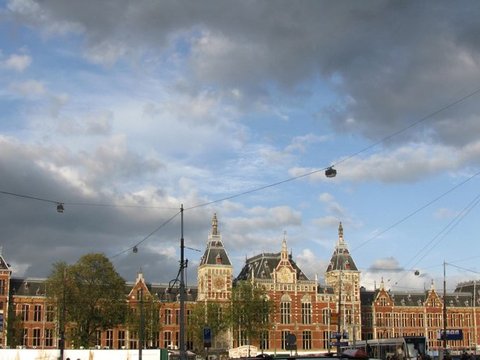 Heavy grey cloud banks over the clock towers of Amsterdam Centraal, the brick facade catching a patch of low sunlight beneath a web of tram wires.