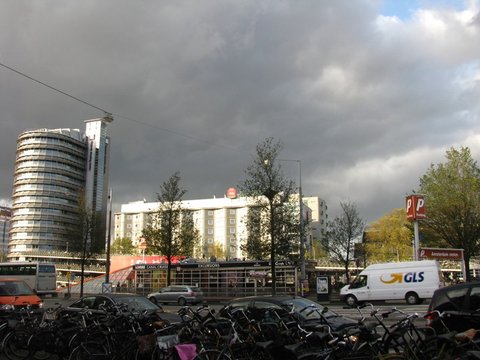 A thicket of parked bicycles in front of a canal-cruise kiosk and a white delivery van, dark storm clouds massing above a curved glass tower.