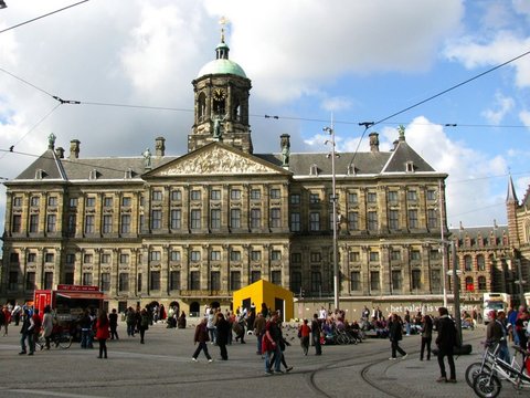 Sunlit facade of the Royal Palace behind a busy square, with a yellow kiosk, a red snack cart, tram tracks and people strolling across the cobbles.