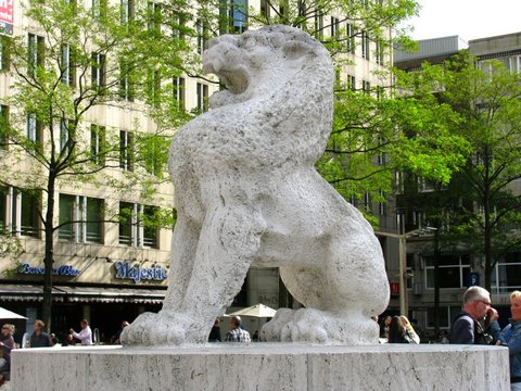 A rough-hewn white stone lion crouches on its plinth, mouth open, against fresh green trees and a tall pale office building.