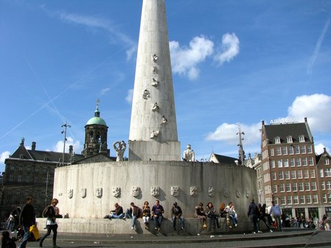 The white travertine pillar of the National Monument rises from a curved wall of carved shields where people sit in the sun, the palace dome behind.