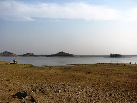 A small boat rests at the edge of a wide, still reservoir, with low wooded islands on the far side and stony ground in the foreground.